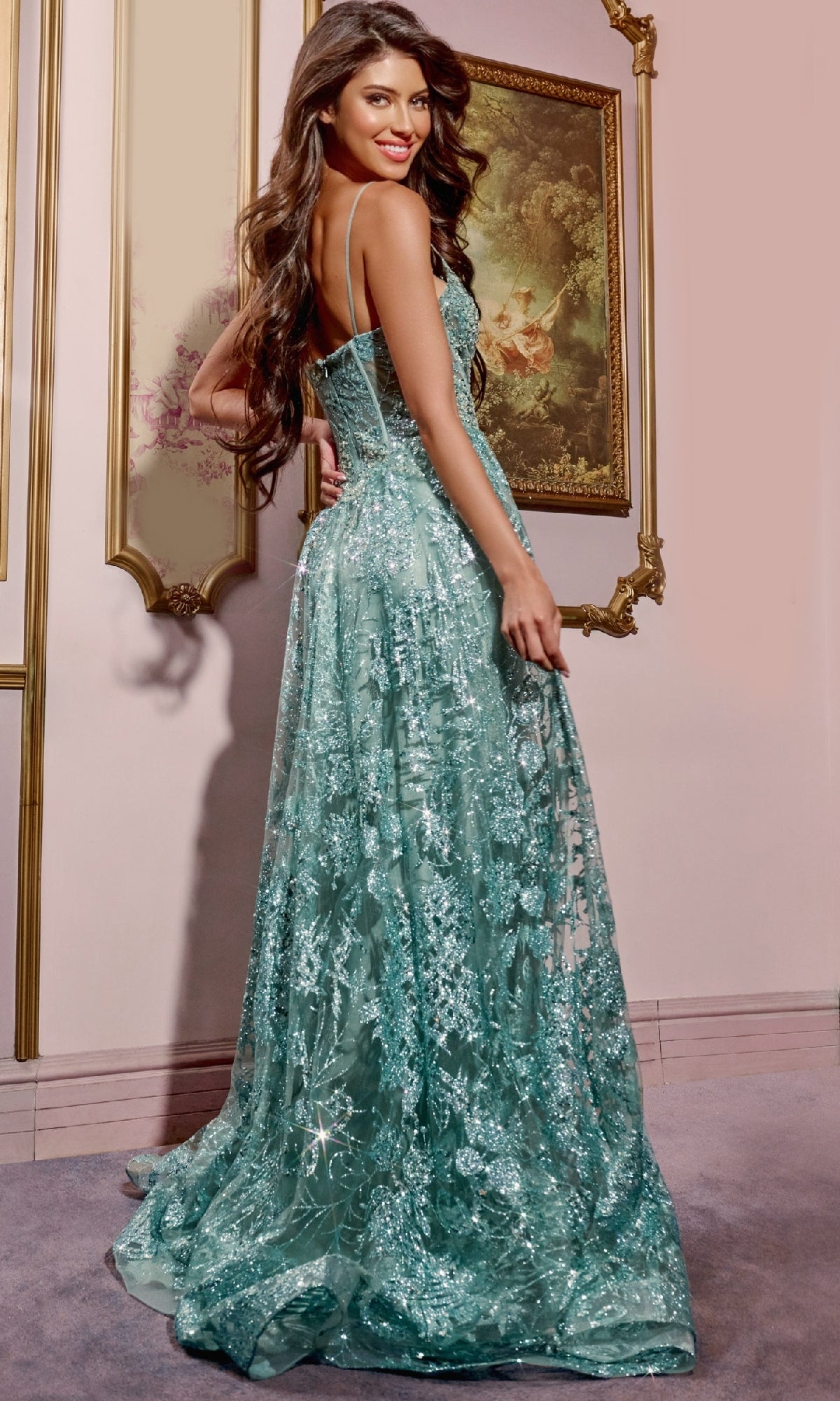A woman in a sparkly teal Jovani 42199 Long A-Line Glitter Prom Dress with a sheer bodice and thin straps stands indoors by ornate framed paintings, looking over her shoulder and smiling.